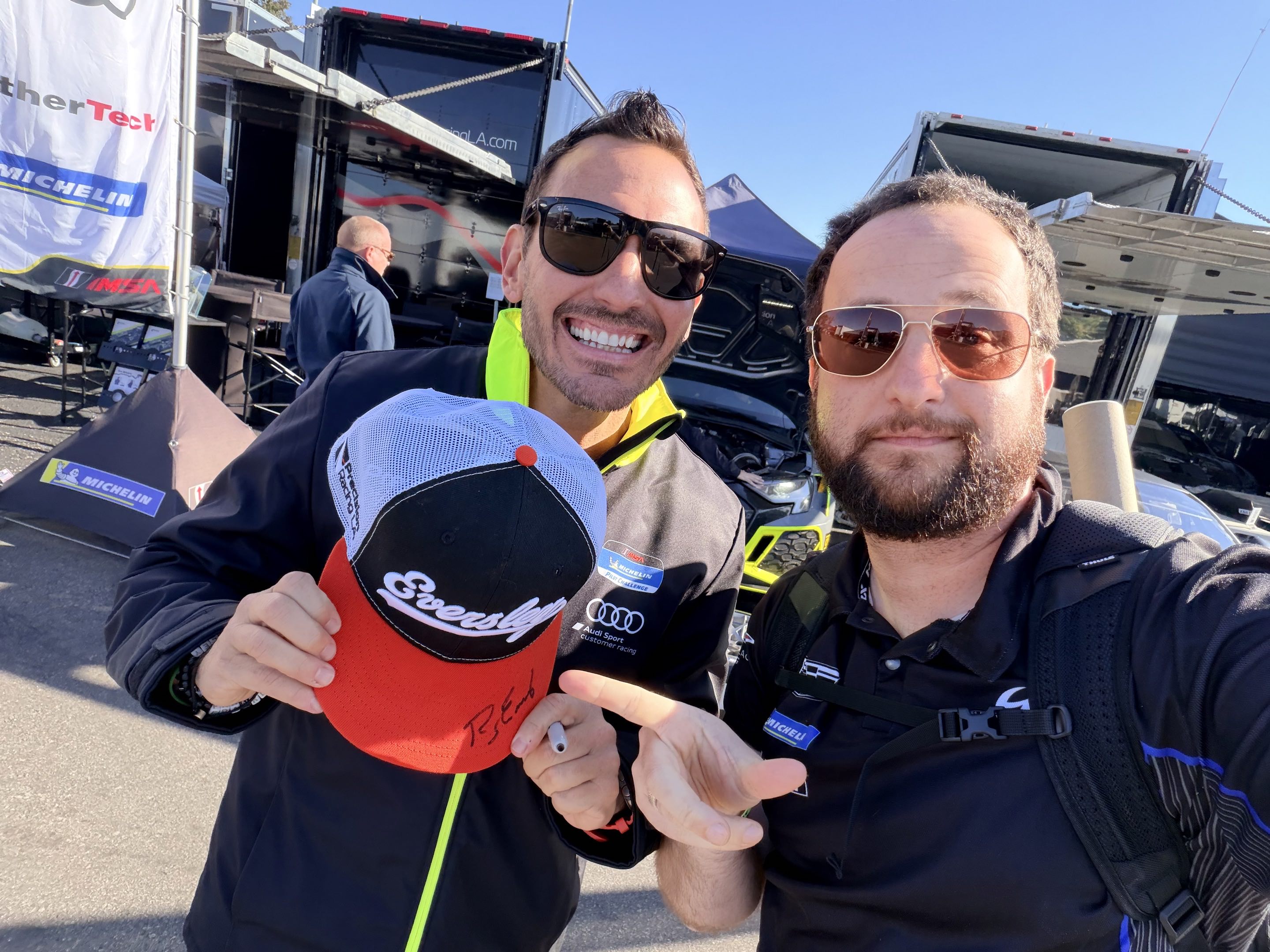 Two men in sunglasses posing with a hat that has been autographed